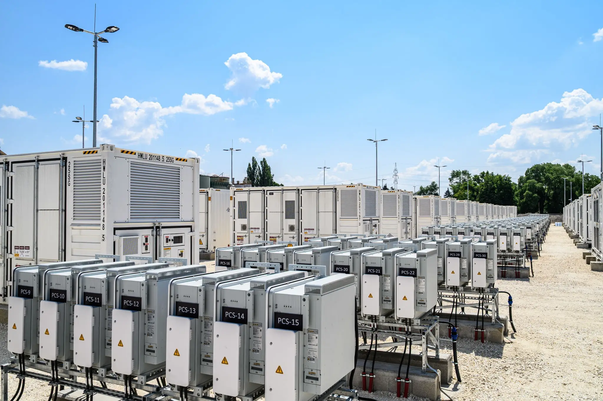 Rows of utility-scale battery energy storage units and power conversion systems are arranged in a large outdoor energy facility under a clear blue sky. White containerized battery modules and electrical cabinets are installed on concrete foundations and connected by cables and equipment. The grid-like layout extends into the distance, illustrating modern energy storage infrastructure that supports renewable energy integration, grid stability, and electricity supply management.