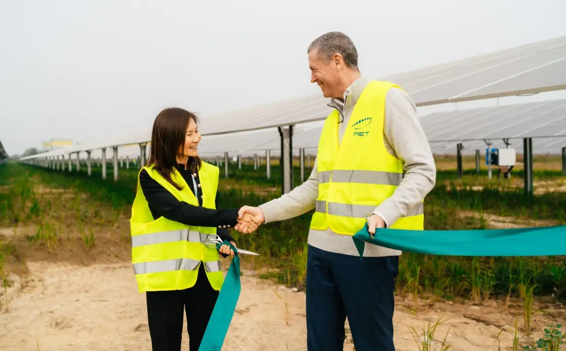 Two people wearing high-visibility yellow safety vests stand at a solar power facility and shake hands while holding sections of a ceremonial ribbon. One person is holding a pair of scissors, suggesting a ribbon-cutting event marking the opening or completion of the site. Rows of solar panels extend into the background across the field, highlighting the renewable energy setting.